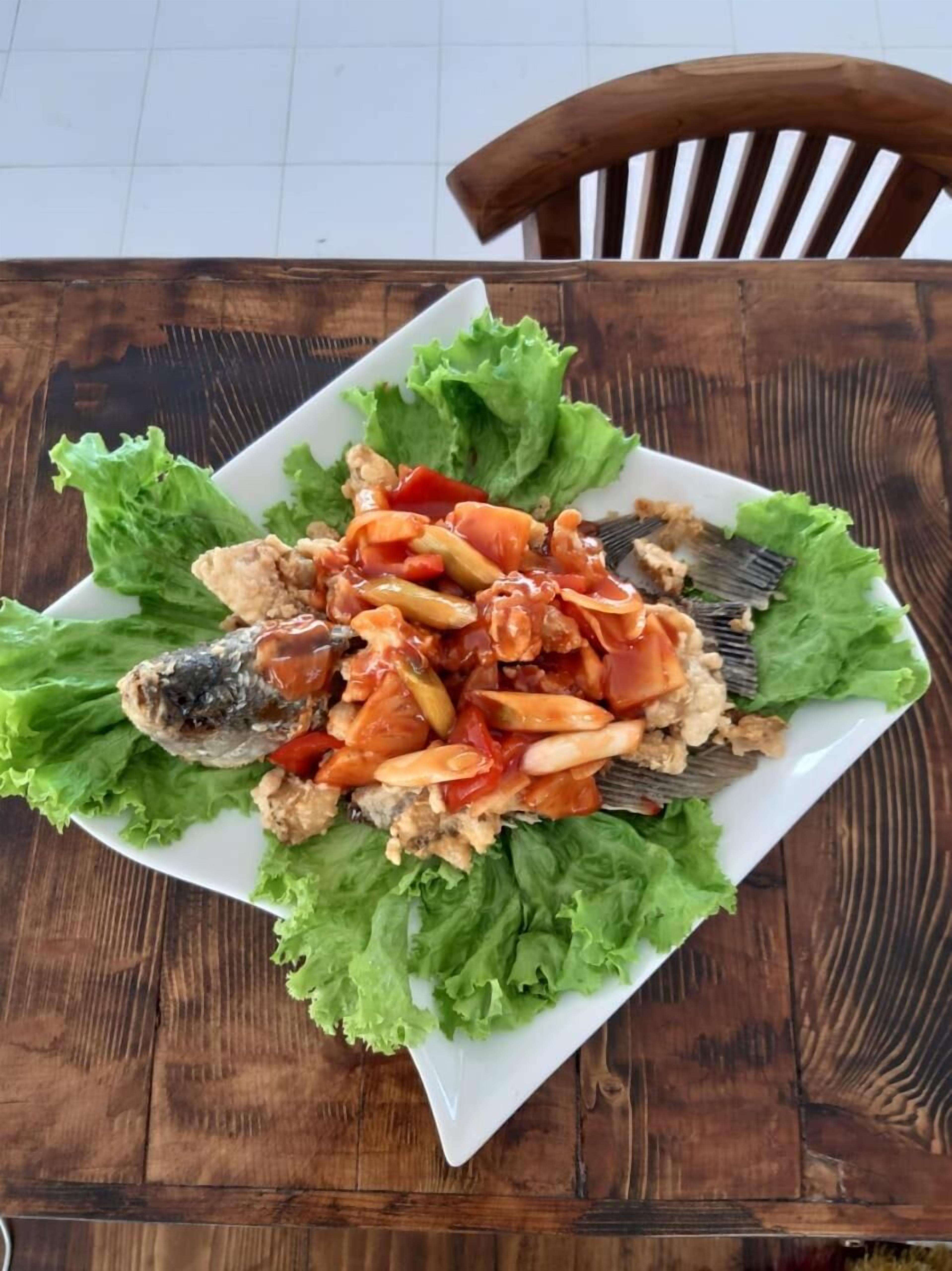 A square white plate with a fish, crab, and vegetables on a wooden table.