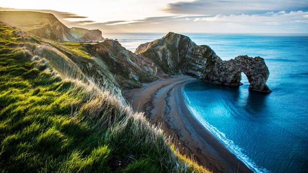 Paisagem costeira com um arco natural de rochas, encostas gramadas e uma praia que desagua no mar.