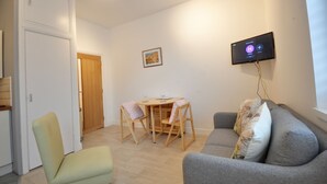 Ein Wohnzimmer mit grauem Sofa, einem kleinen Tisch mit Stühlen, einem an der Wand befestigten Fernseher und einer Holztür.