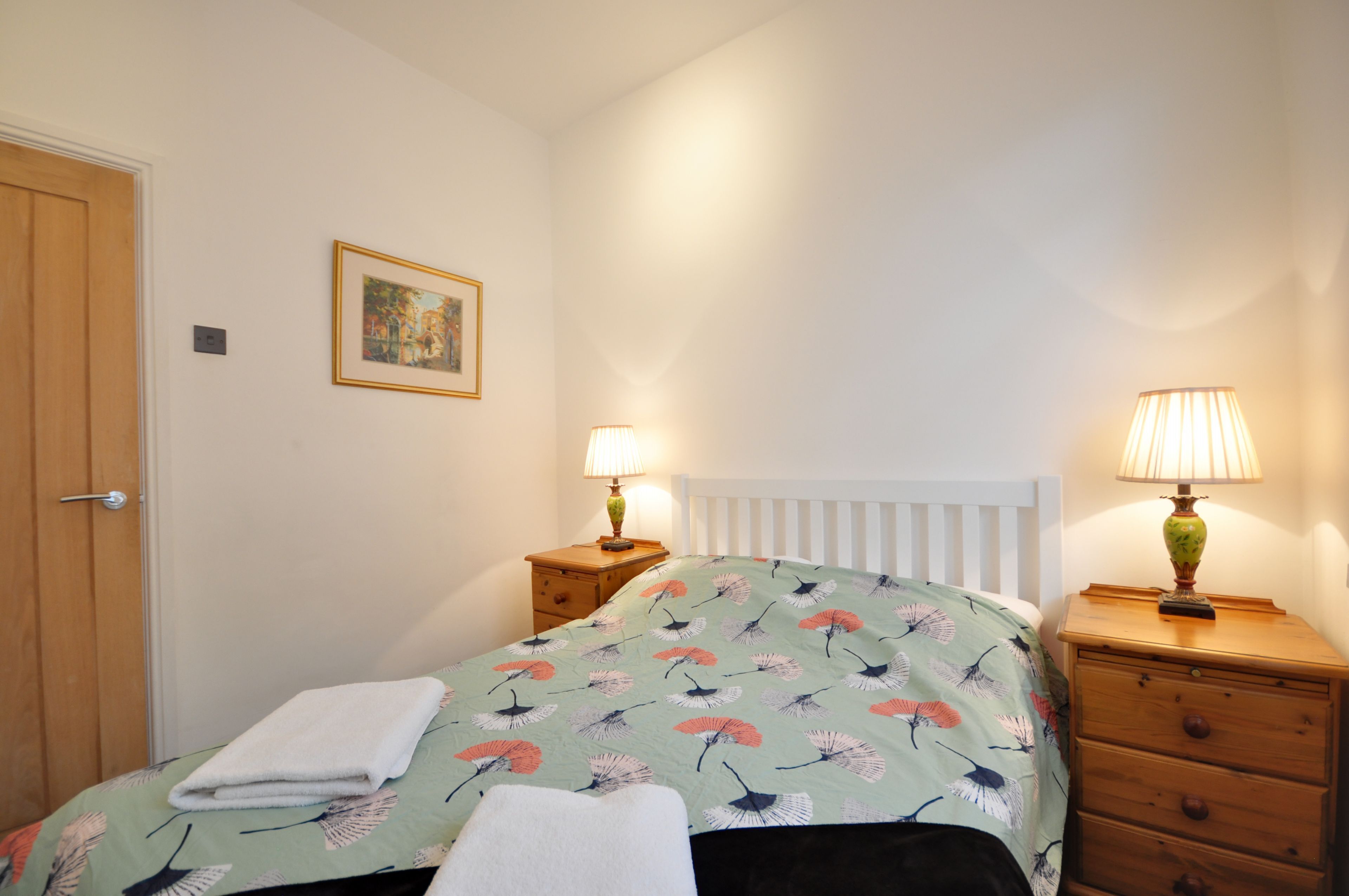 Une chambre à coucher avec un lit, deux tables de chevet avec des lampes, une porte en bois, un tableau encadré au mur et un radiateur blanc.