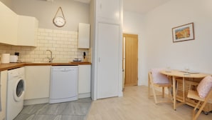 Eine kompakte Küche mit Geschirrspüler, einem weißen Kühlschrank, einem Holzessttisch mit Stühlen und einer Wanduhr.