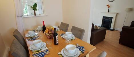 A dining area with a wooden table set for four, surrounded by grey chairs. A bottle of wine and a fruit bowl are on the table.
