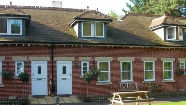 Un bâtiment en briques rouges, doté de fenêtres à cadre blanc, d’une porte d’entrée et d’un petit jardin avec une table de pique-nique.