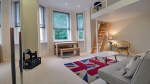Un salon moderne avec une chambre à l’étage, accessible par un escalier, comprenant un canapé, une petite table et un tapis coloré.