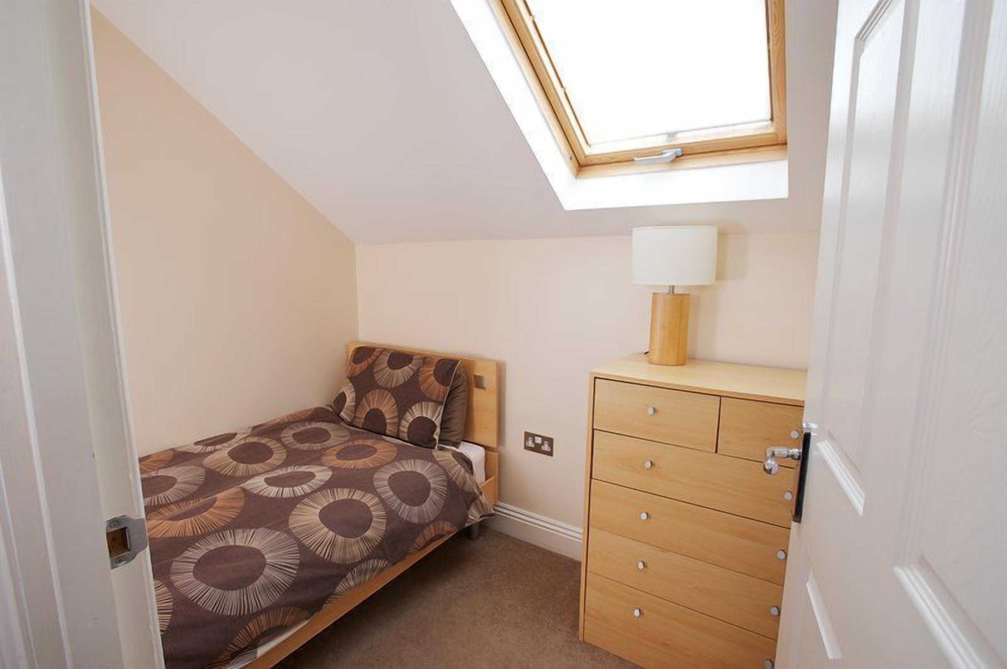 A bedroom with a skylight, a bed with a patterned bedspread, a bedside table with a lamp, and a wooden dresser.