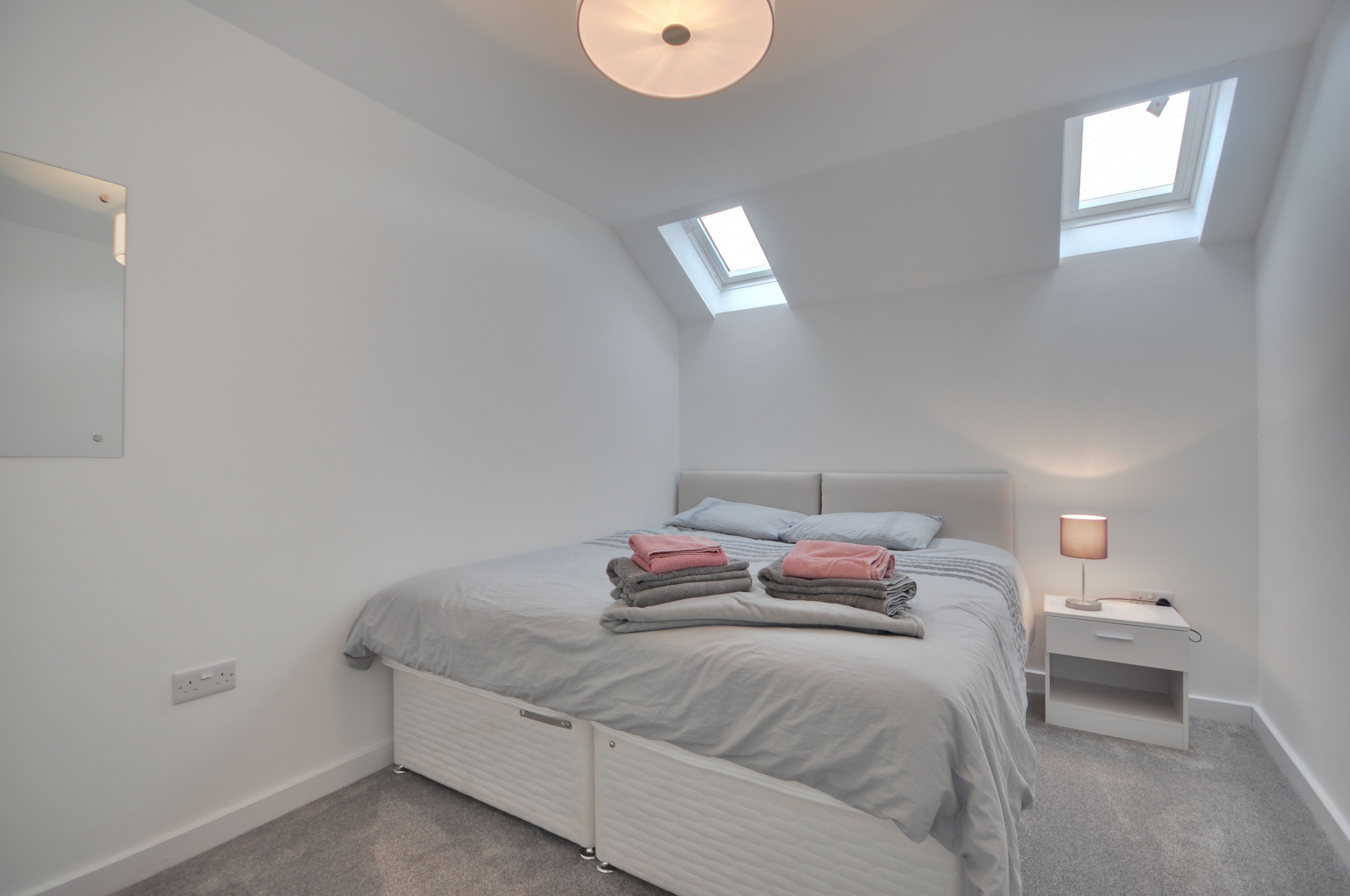 Une chambre à coucher comprenant un lit, deux tables de chevet, une petite table, un puits de lumière et un luminaire fixé au mur.
