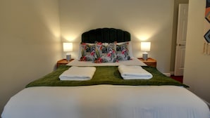 Un lit bien fait, avec une couvre-lit verte et deux serviettes blanches, un ensemble d’oreillers à motifs floraux et deux lampes de chevet posées sur des tables de nuit en bois.
