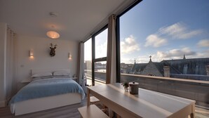 Un dormitorio con un ventanal grande, una cama, un banco de madera y una mesa con un florero.