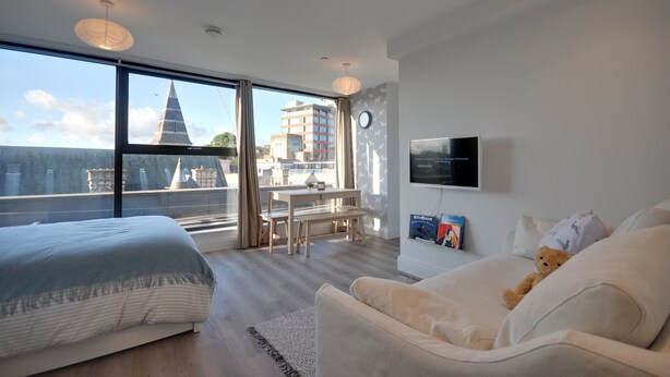 Una habitación de hotel moderna con un ventanal grande, una cama, un sofá, un televisor y una mesita.