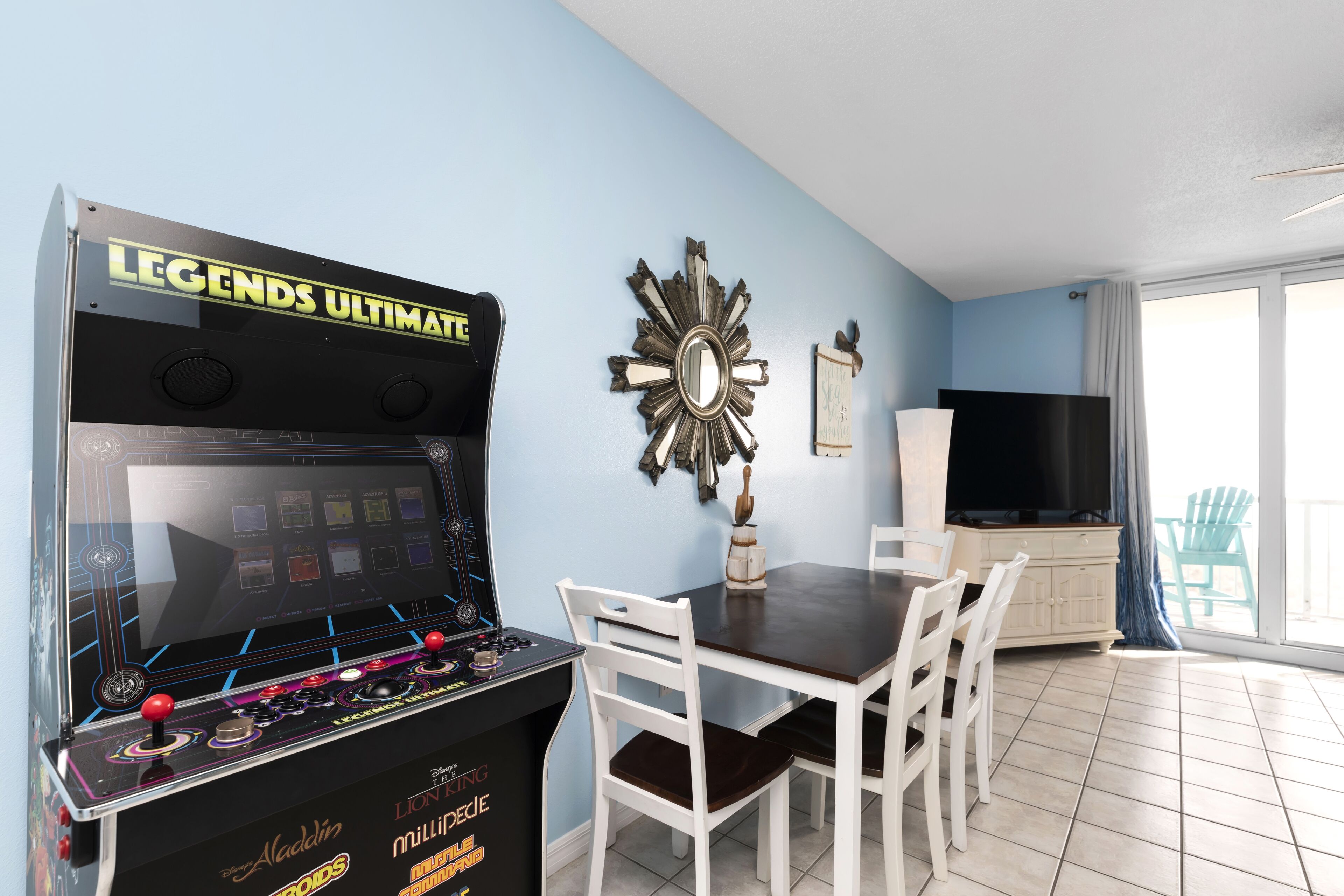 Een kamer met een spelcomputer, een eettafel met stoelen, een televisie en een balkon met een stoel.