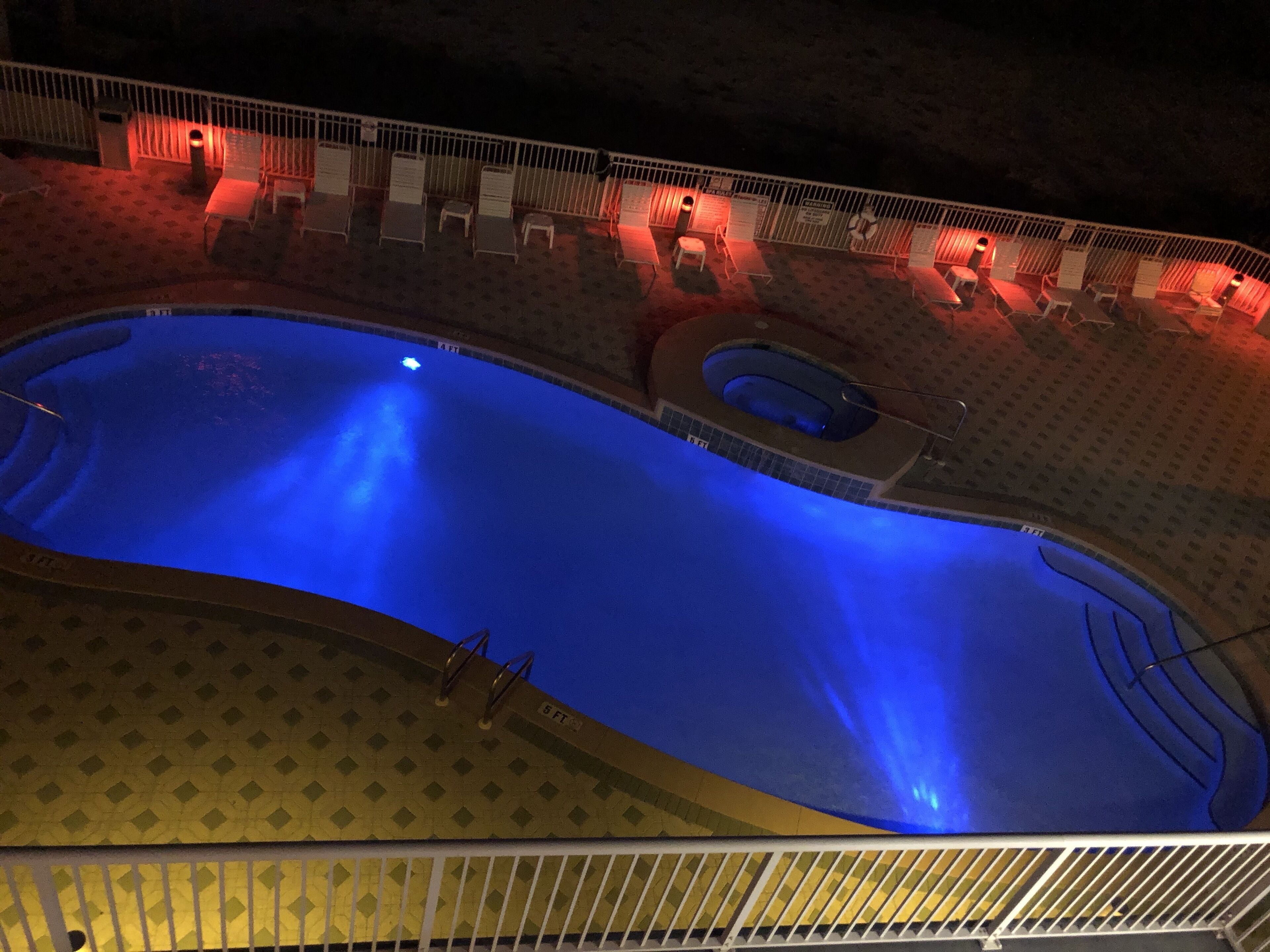 Ein Schwimmbecken mit Sprungbrett, umgeben von Liegestühlen und einer Liegebereich, mit Zaun und Schildern mit Regeln.
