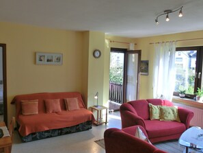 Un salon avec un canapé rouge, un petit fauteuil rouge et une table basse en verre. On y trouve une petite table d’appoint avec une lampe, ainsi qu’une fenêtre avec des rideaux.