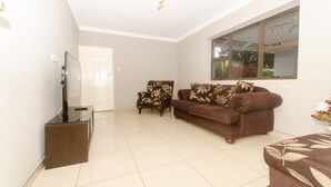 A living room with a brown sofa, a chair, a TV stand, and a window with a view of greenery. - Sarum Lodge (Harare)