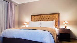 A hotel room with a tufted headboard, a large bed with white linens, two bedside tables with lamps, and a window with curtains. - Sarum Lodge (Harare)
