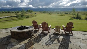 A patio with Adirondack chairs and a stone fire pit overlooking a grassy field and mountains. - Bunchberry Court Luxury Home at Tributary in Driggs, Idaho with Amazing Views (Driggs)