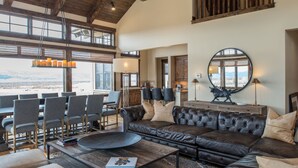 A spacious living room with a high ceiling, wooden beams, a large window, a leather sofa, and a dining area with chairs. - Bunchberry Court Luxury Home at Tributary in Driggs, Idaho with Amazing Views (Driggs)