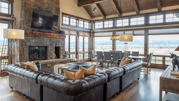 A spacious living room with a stone fireplace, leather sofa, and large windows offering mountain views. - Bunchberry Court Luxury Home at Tributary in Driggs, Idaho with Amazing Views (Driggs)