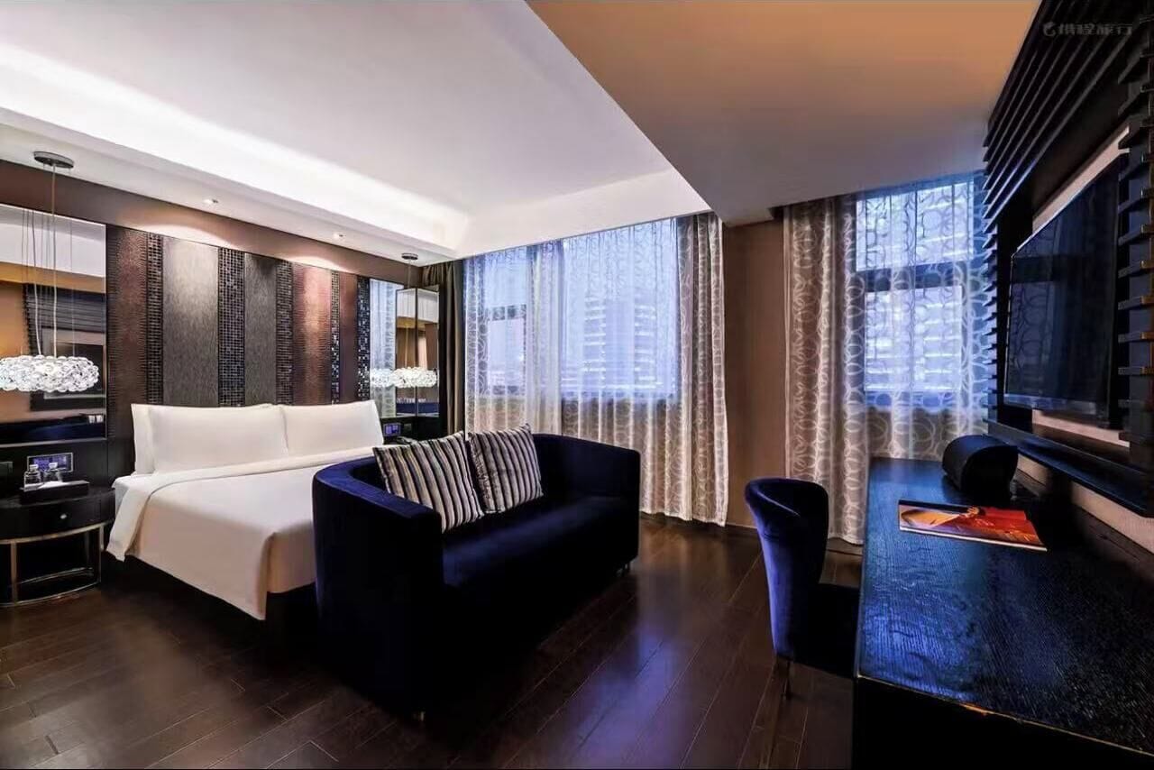 Una habitación de hotel moderna con una cama grande, un sofá de color oscuro, una mesita y un televisor.