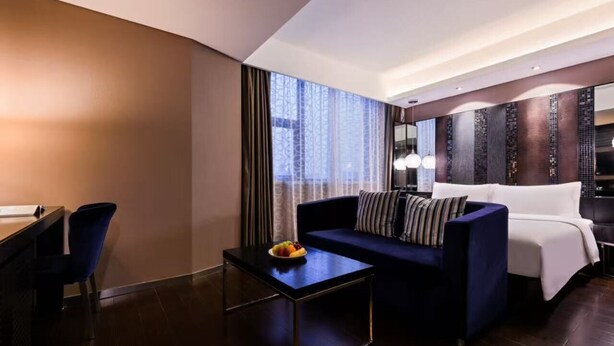 A modern hotel room with a dark wood floor, a sofa, a small table with fruit, a desk, and a large window with curtains.