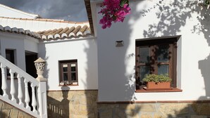 Un edificio blanco con fachada de piedra, un patio con tejas, una escalera de madera y una ventana con planta en maceta.
