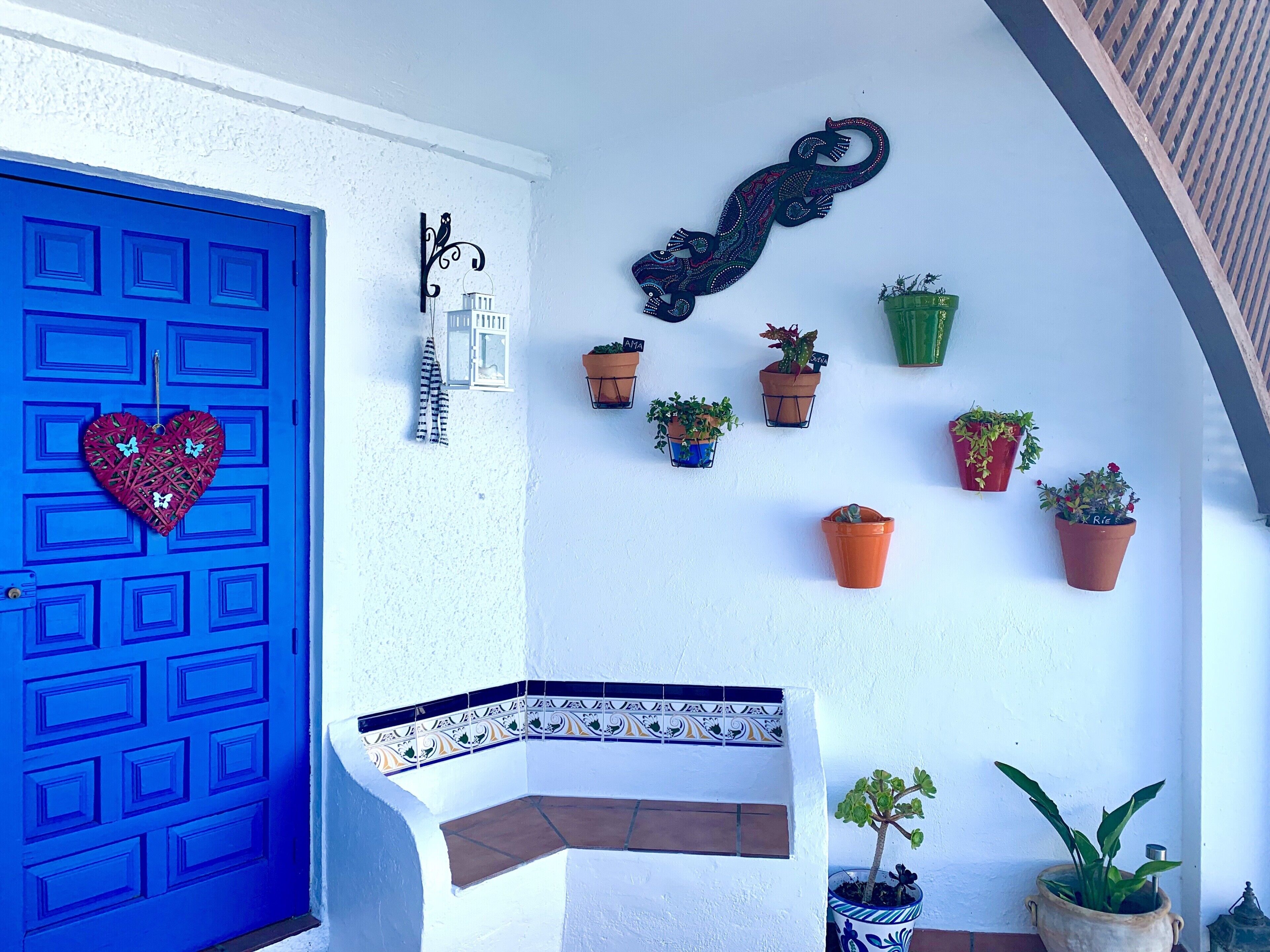 A blue door with a heart decoration, a white wall with a black decorative piece, and several potted plants arranged on the wall.