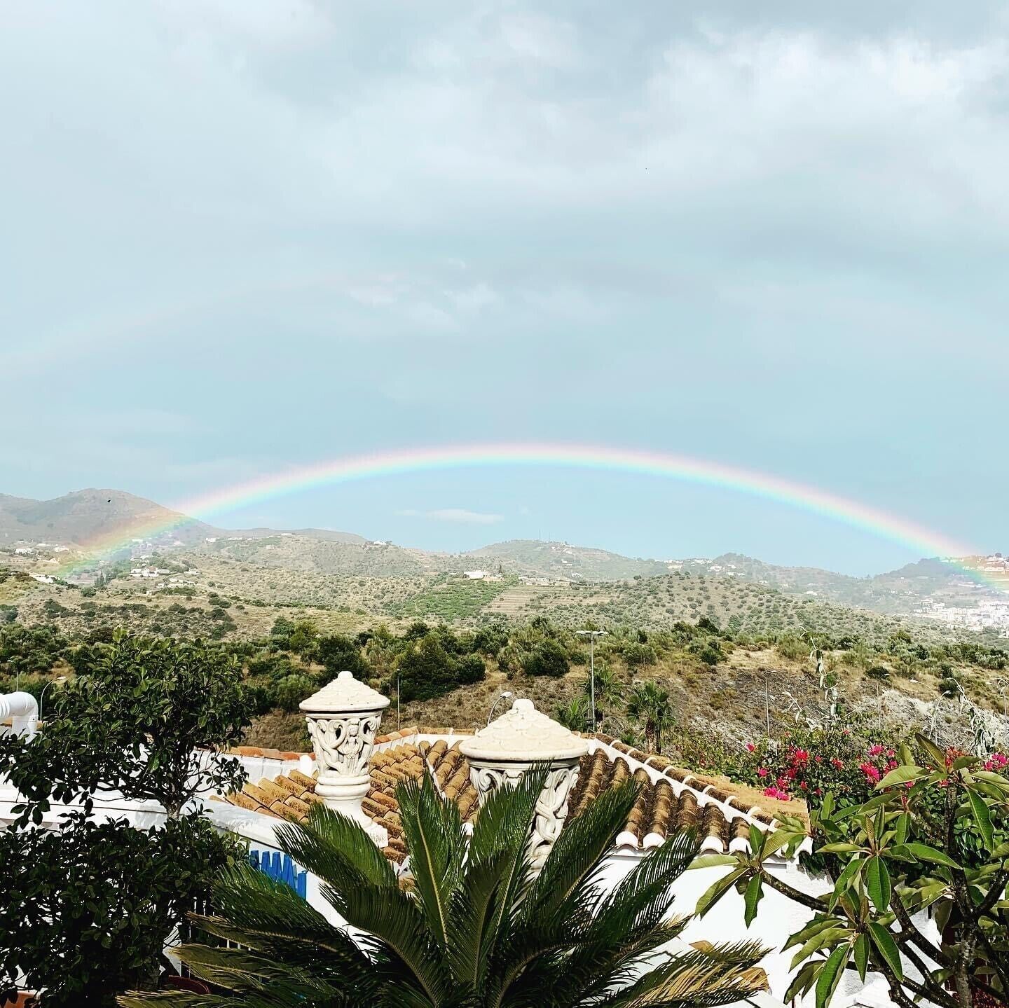 Panorama montuoso con un arcobaleno, che include un edificio bianco con tetto in tegole e una colonna decorativa.