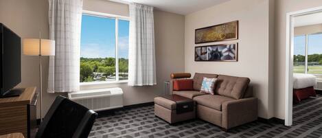 Quarto de hotel com sofá, televisão, escrivaninha e janela com cortinas.