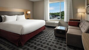 A hotel room with a large bed, a sofa, a small table, and a window with a view of greenery and buildings. - TownePlace Suites by Marriott Memphis Olive Branch (Olive Branch)