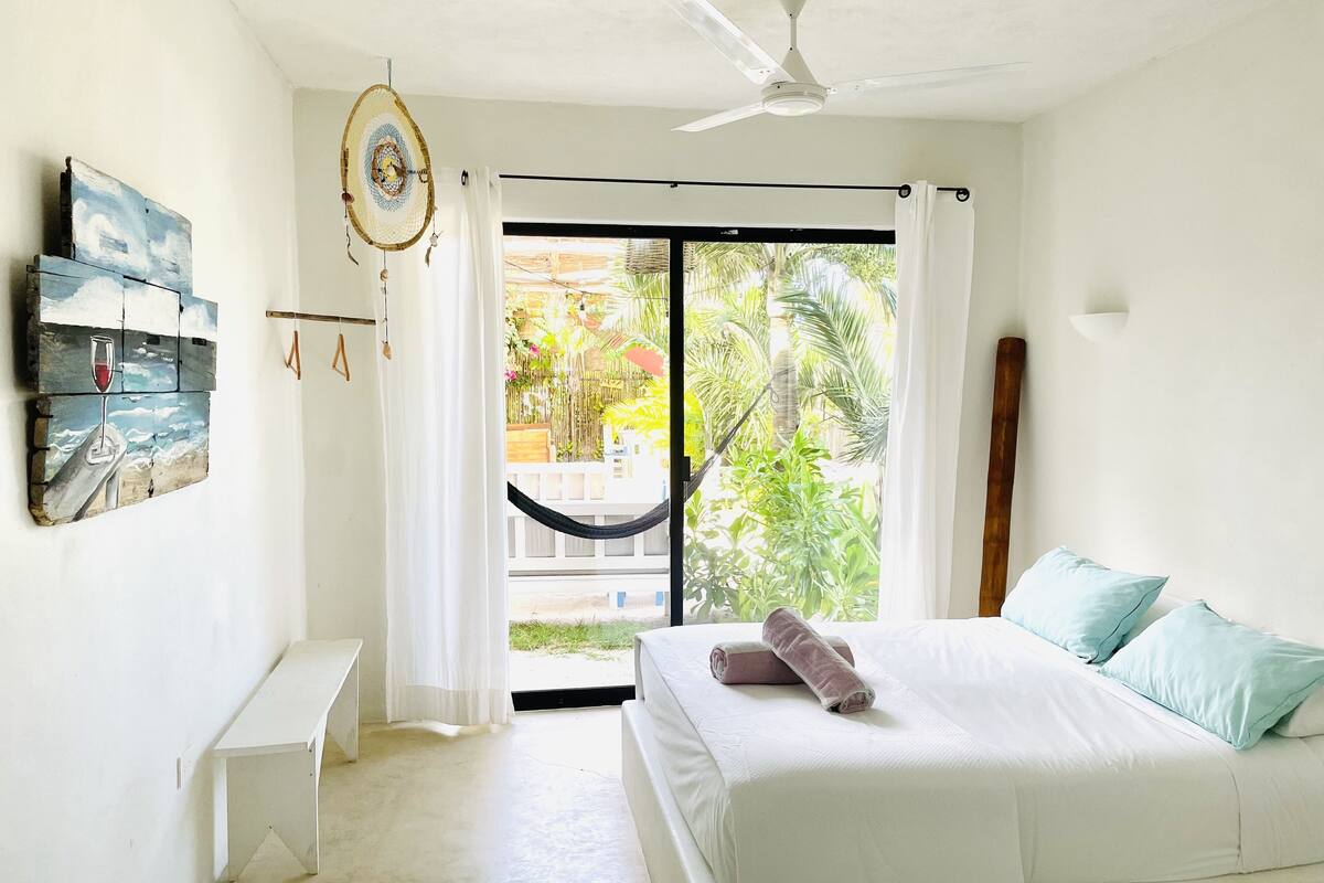 A bedroom with a bed, a bench, a ceiling fan, and a view of a hammock and greenery outside.