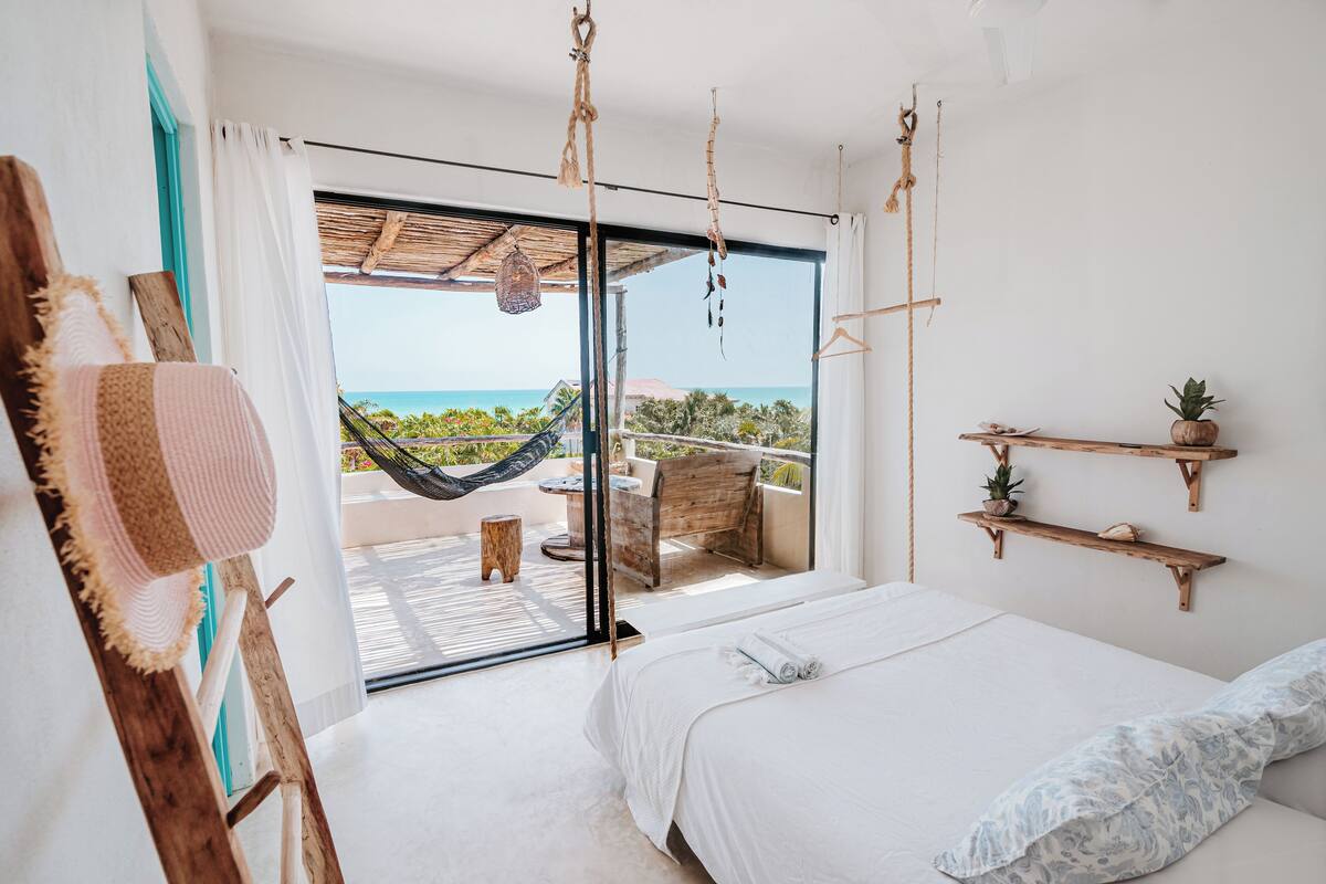 A bedroom with a large bed, a hammock, a wooden shelf with plants, and a view of the ocean.