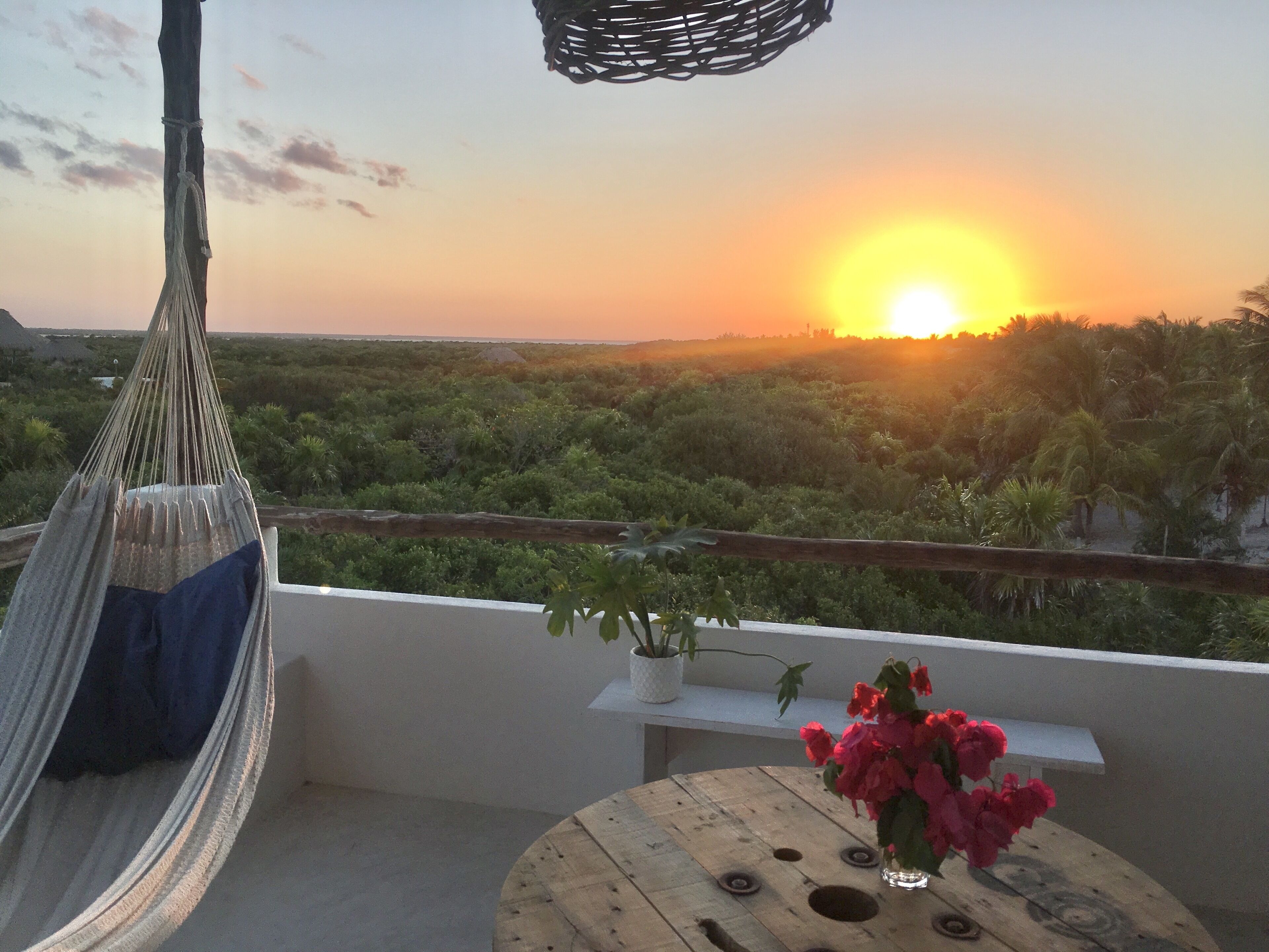 A hammock chair with a cushion, a small table with a vase of flowers, and a view of a sunset over a landscape with trees.