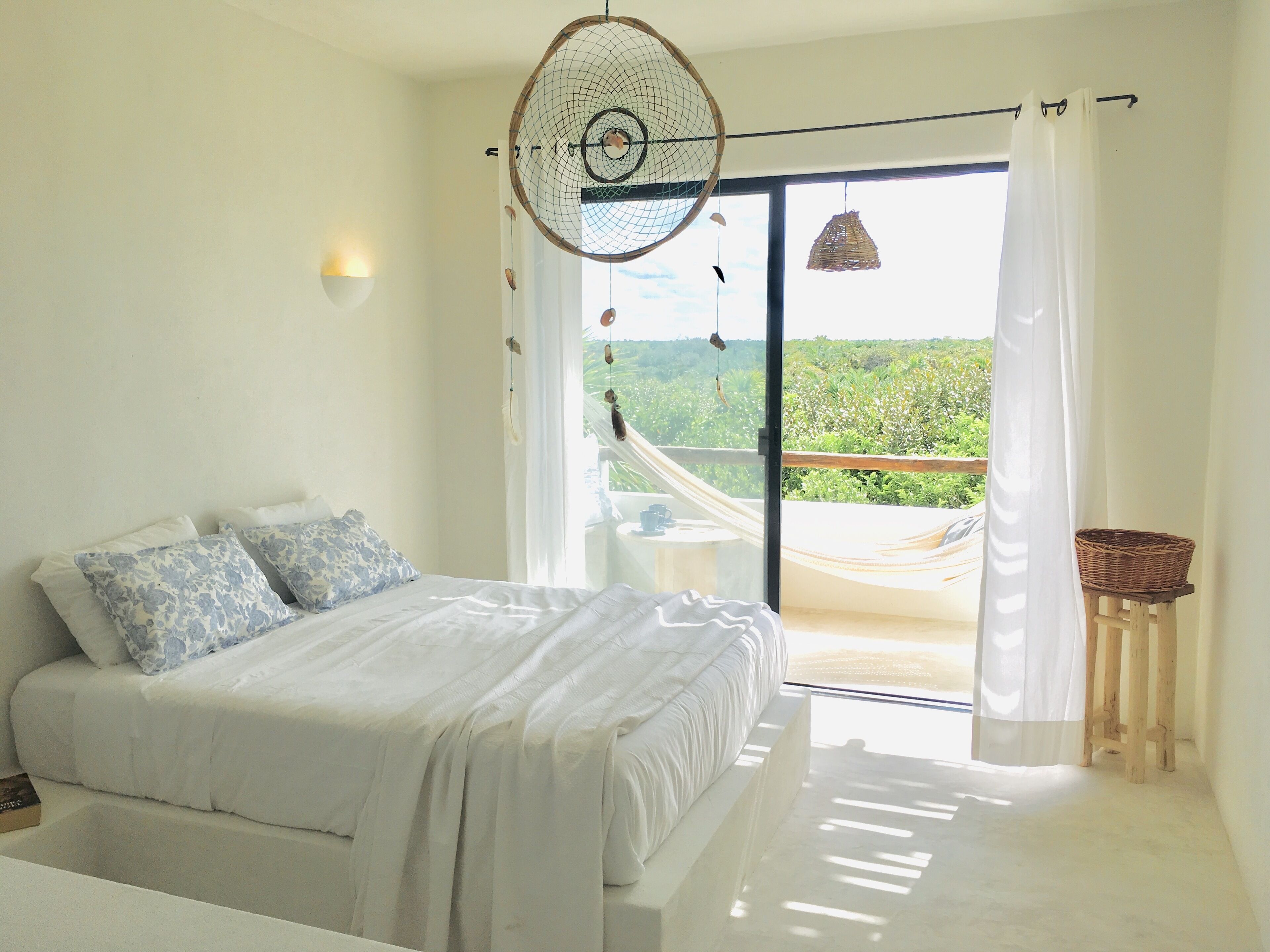 A bedroom with a bed, a nightstand, a hammock, and a view of greenery outside.