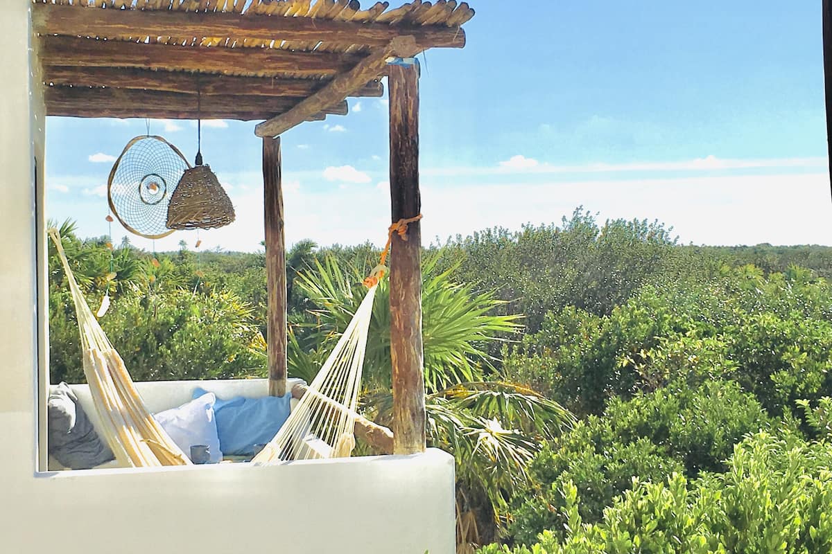 A hammock under a wooden pergola with a hanging lantern, overlooking a lush green landscape.