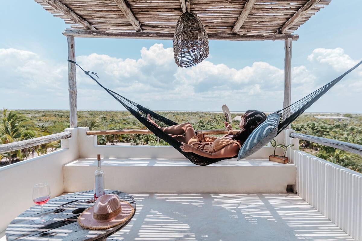 A hammock under a wooden pergola on a balcony with a table, a glass of wine, and a hat.