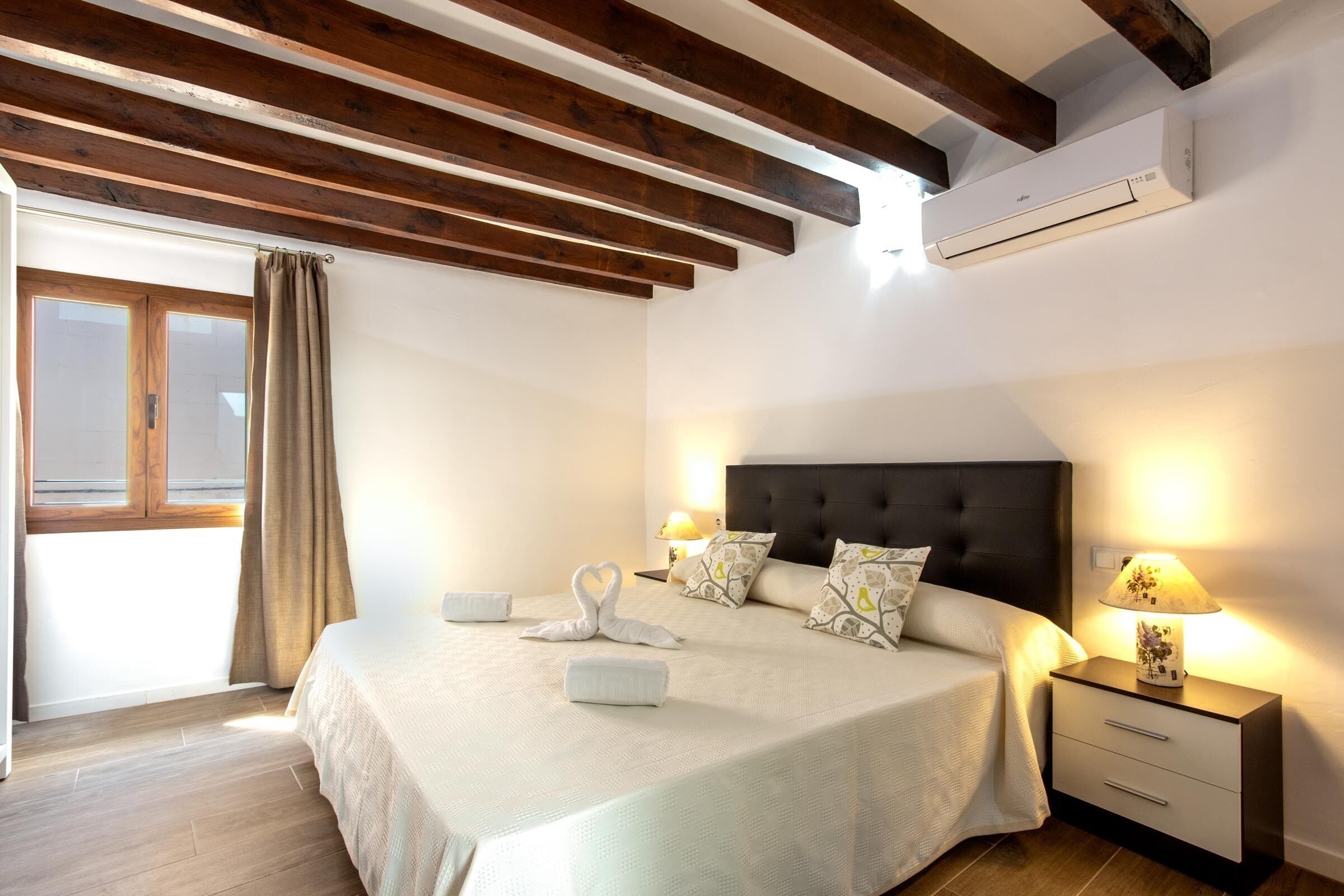 A bedroom with a bed, bedside tables, a window with curtains, wooden beams, and an air conditioning unit.