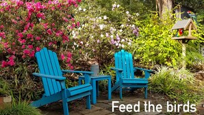 A garden with two blue Adirondack chairs, a bird feeder, and a variety of flowering plants.