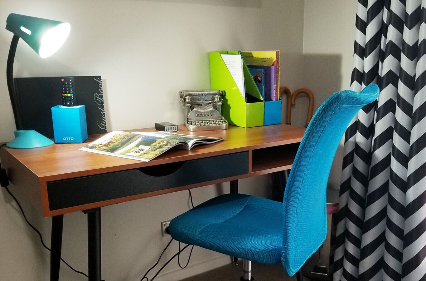 A desk with a lamp, a book, a Corona typewriter, and a chair.