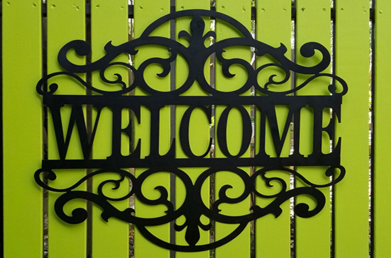 A black metal sign with the word "WELCOME" in capital letters, mounted on a green background with vertical lines.