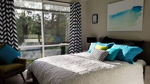 A bedroom with a large bed, a view of trees through a window, and a painting on the wall.