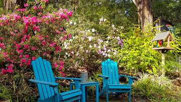 A garden with two blue Adirondack chairs, a bird feeder, and a variety of flowering plants.