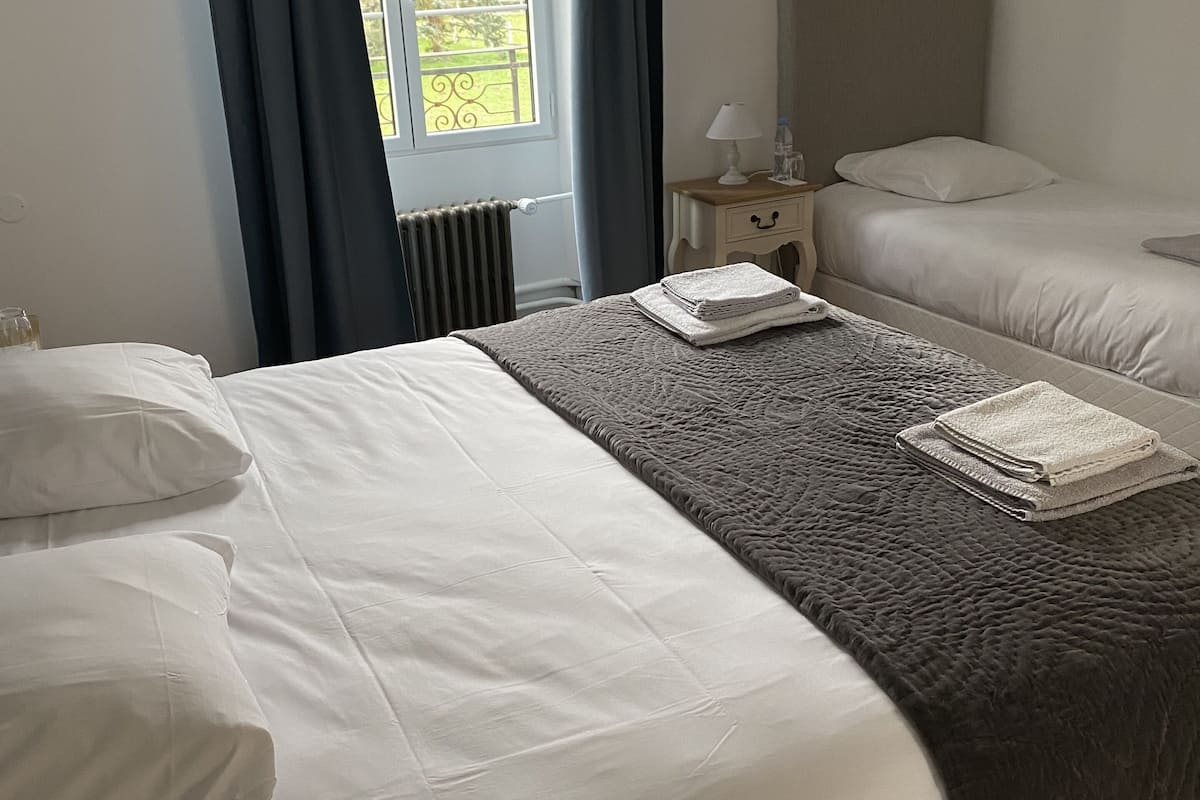 A hotel room with two beds, a window with curtains, a bedside table with a lamp, and a view of greenery outside.