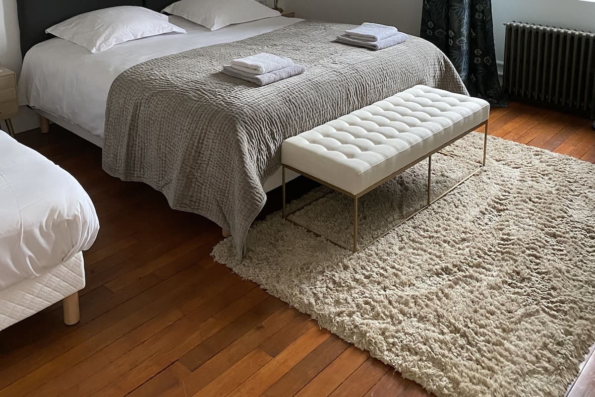 A bedroom with a bed, bedside tables, a bench, a window with curtains, and a radiator.