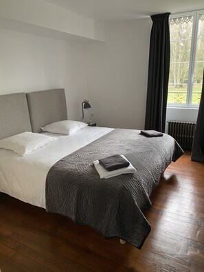 Un lit bien fait, avec une tête de lit grise, des oreillers blancs et une couvre-lit matelassée grise. On aperçoit une petite table de chevet avec une lampe et des bouteilles d’eau. Une fenêtre donne sur un paysage verdoyant.