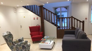 Un salón con un sillón estampado, una silla roja, una mesa de centro de cristal y un sofá gris. Se ve una escalera de madera con barandilla al fondo.