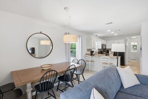 A modern kitchen with white cabinets, a dining area with a wooden table and black chairs, and a living room with a blue sofa.