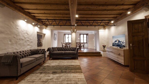 Een ruime woonkamer met een hoog houten plafond, een grote bank, een eethoek en een houten kast.