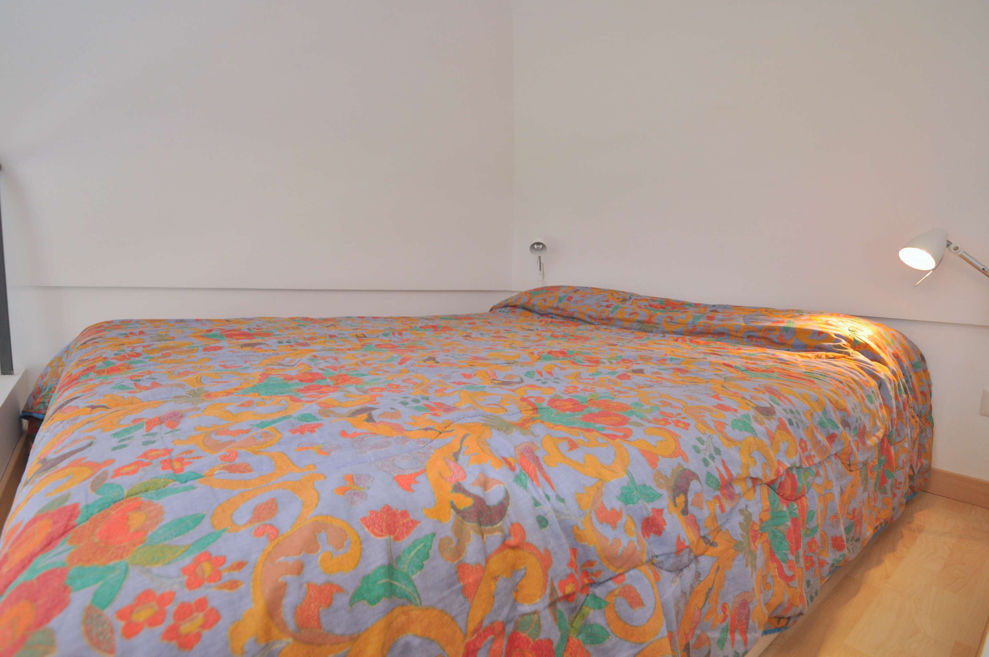 Ein Bett mit farbig-floralem Überwurf, einer Nachttischlampe und einer schlichten Wand.
