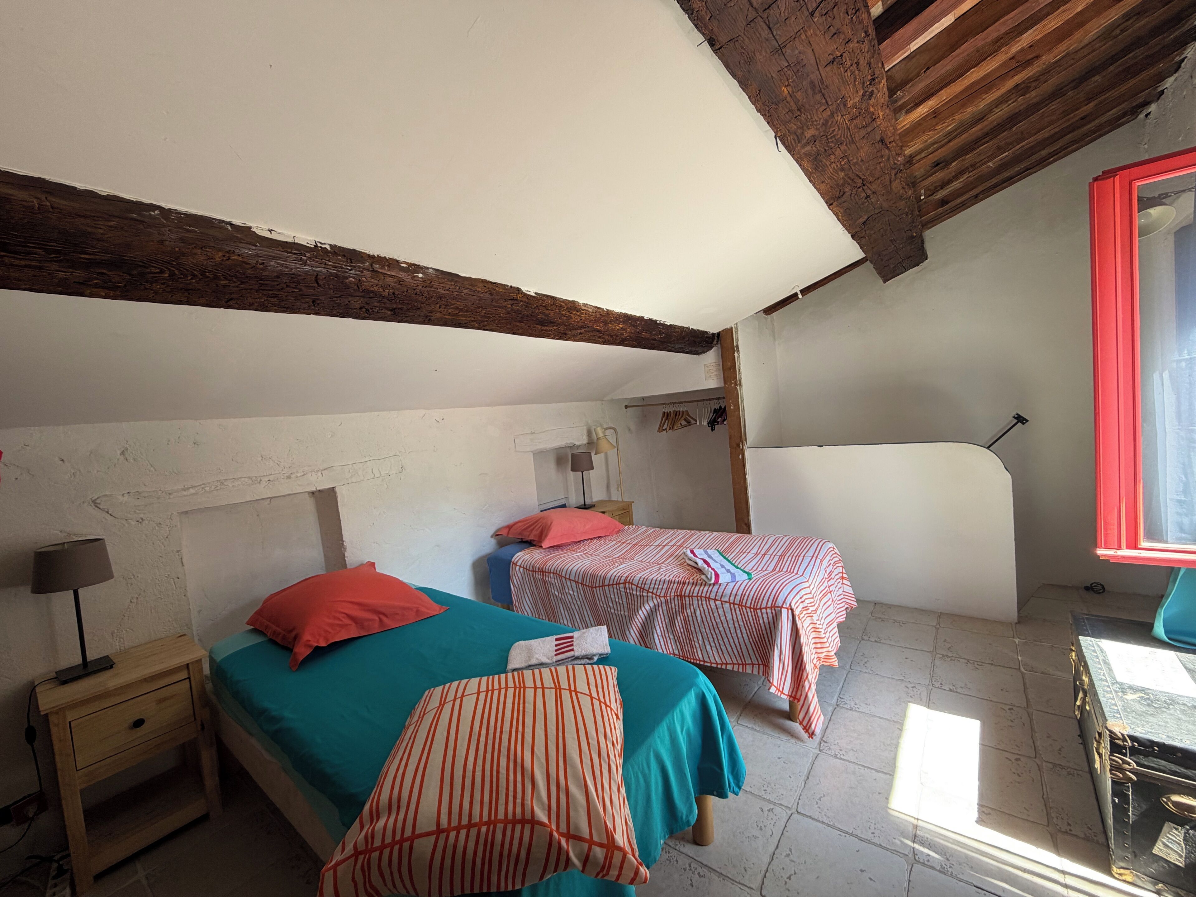A room with two beds, a wooden ceiling, a window with red trim, and a table with a chair.