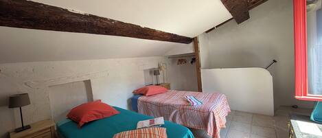 A room with two beds, a wooden ceiling, a window with red trim, and a table with a chair.
