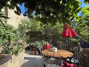 En udendørs terrasse med et rundt træbord, metalstole, en rød parasol og forskellige planter.